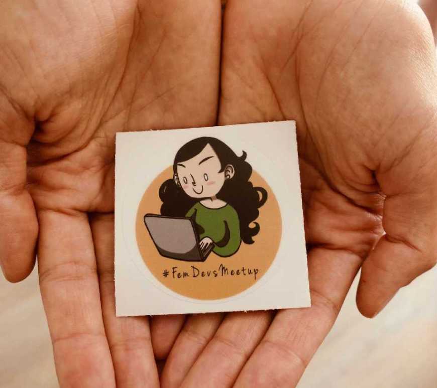 Das Logo des #FemDevsMeetups auf einem Sticker präsentiert in zwei Händen.